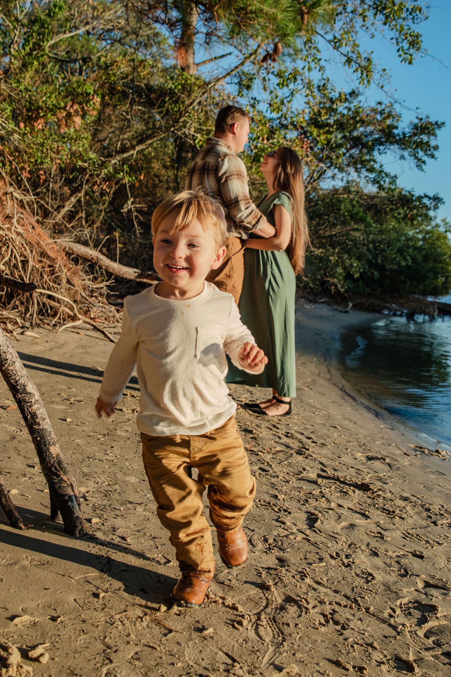 A happy toddler runs on a sandy beach while his parents stand together near lush trees in warm golden light.