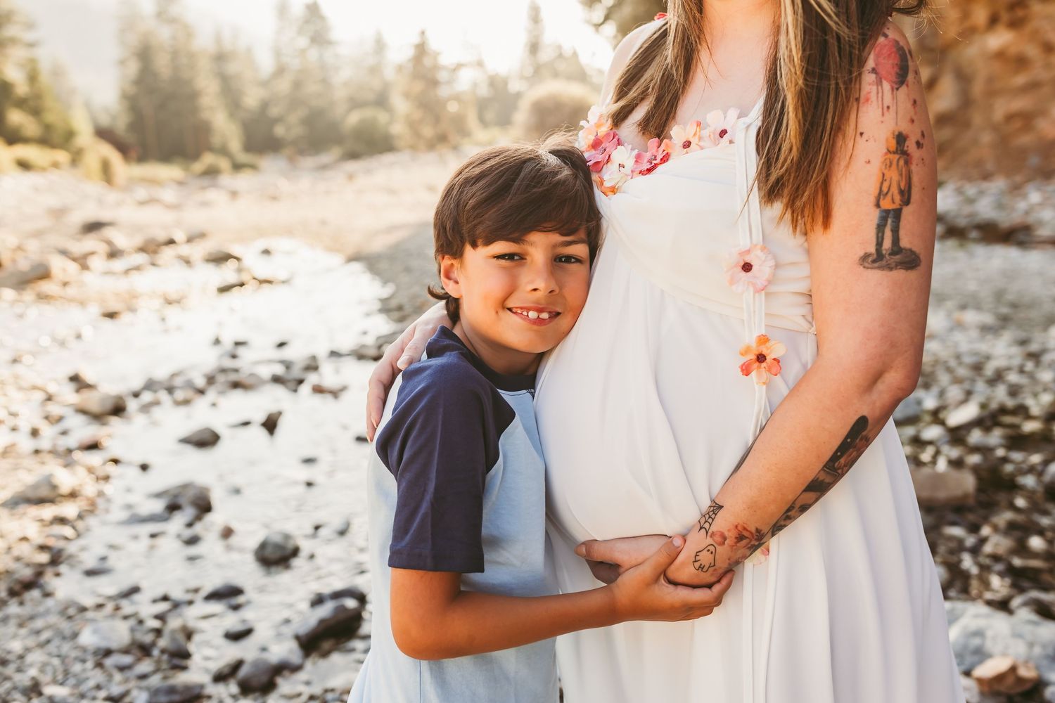 A young boy hugs a pregnant woman in a white dress outdoors near a rocky river, smiling warmly at the camera.