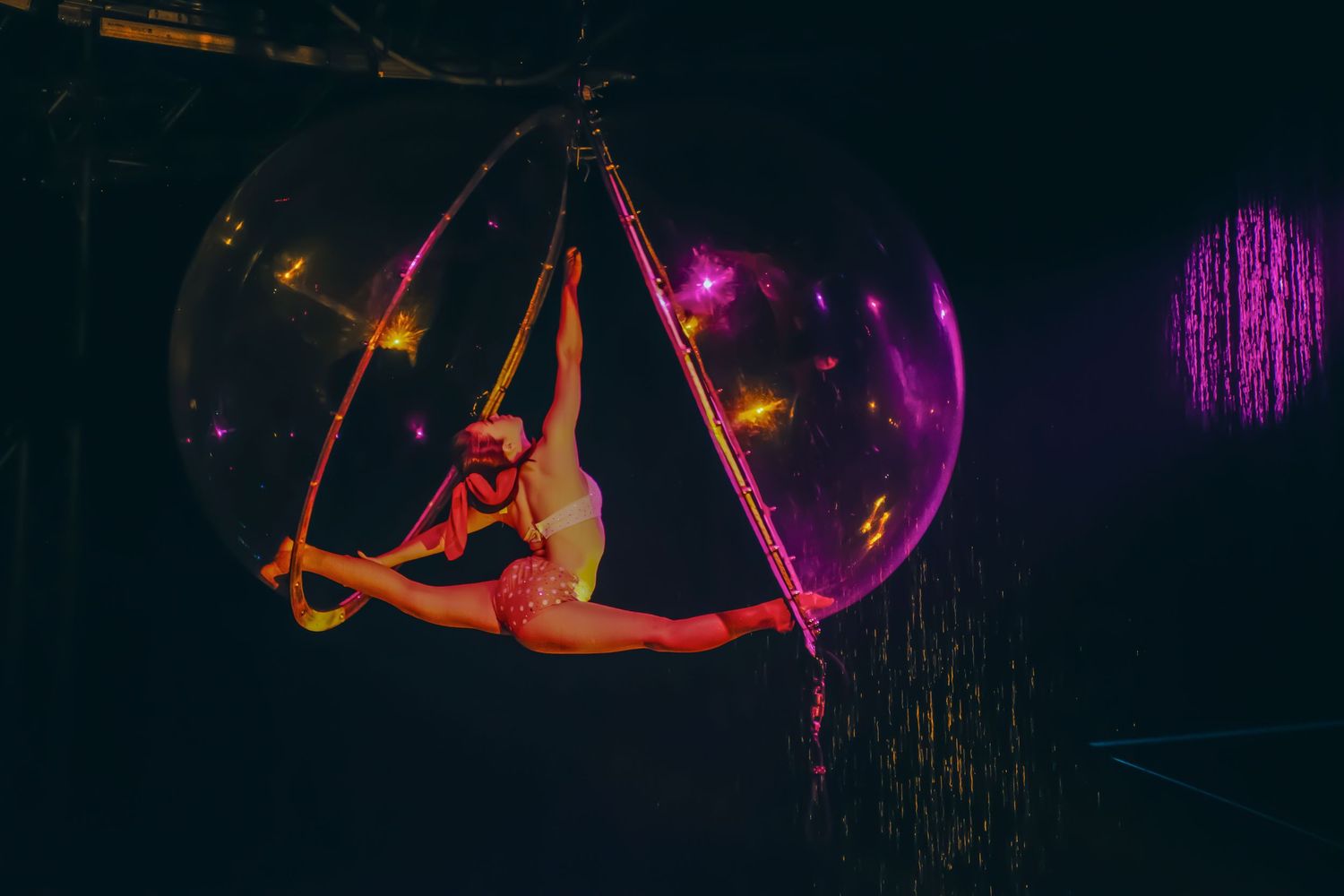 Aerial acrobat performs splits on suspended hoop surrounded by glowing pink bubbles in dramatic circus performance.