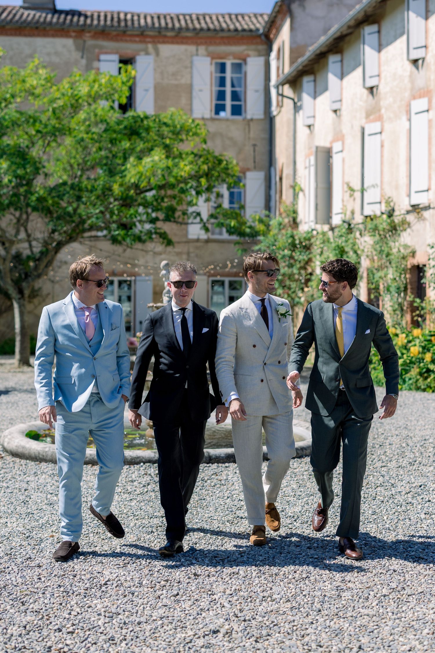 Four stylish men in suits walking confidently outdoors near a rustic French chateau on a sunny day.