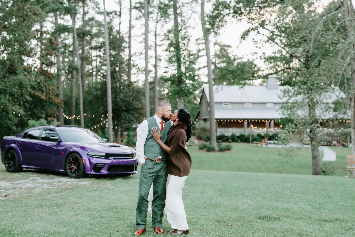 Couple embracing on a green lawn beside a purple sports car with a rustic wooden venue and tall pine trees in the background.