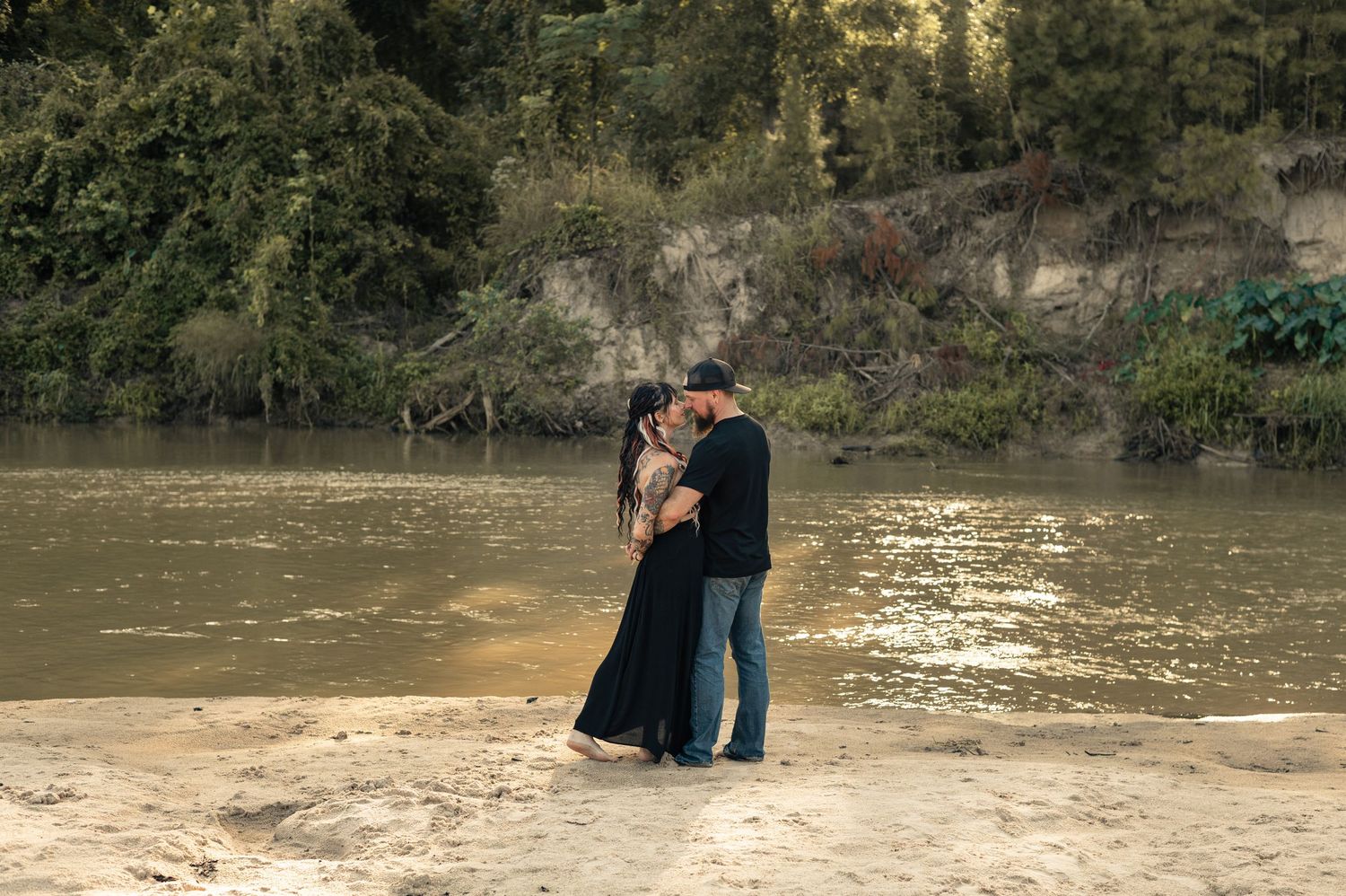 Couple sharing a romantic kiss on a sandy riverbank surrounded by lush green trees and calm water.