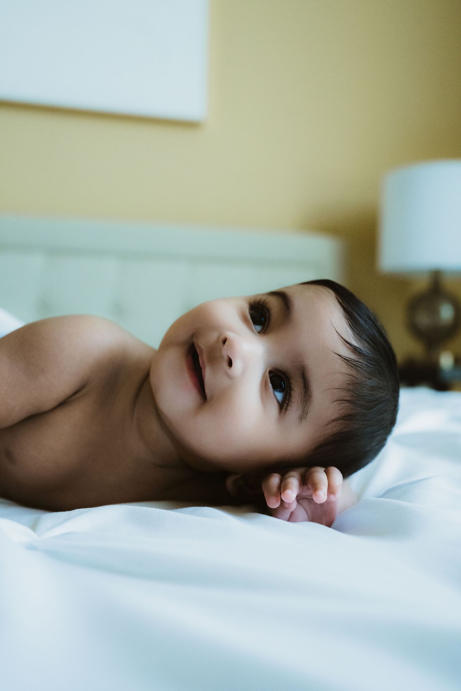 A cute baby with dark hair lying on white bedding, looking up with bright eyes in a softly lit bedroom.