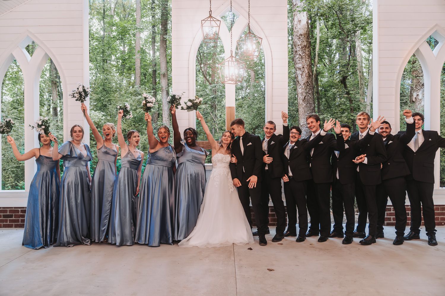 Wedding party posing under white arch with bridesmaids in blue gowns and groomsmen in black suits cheering outdoors.