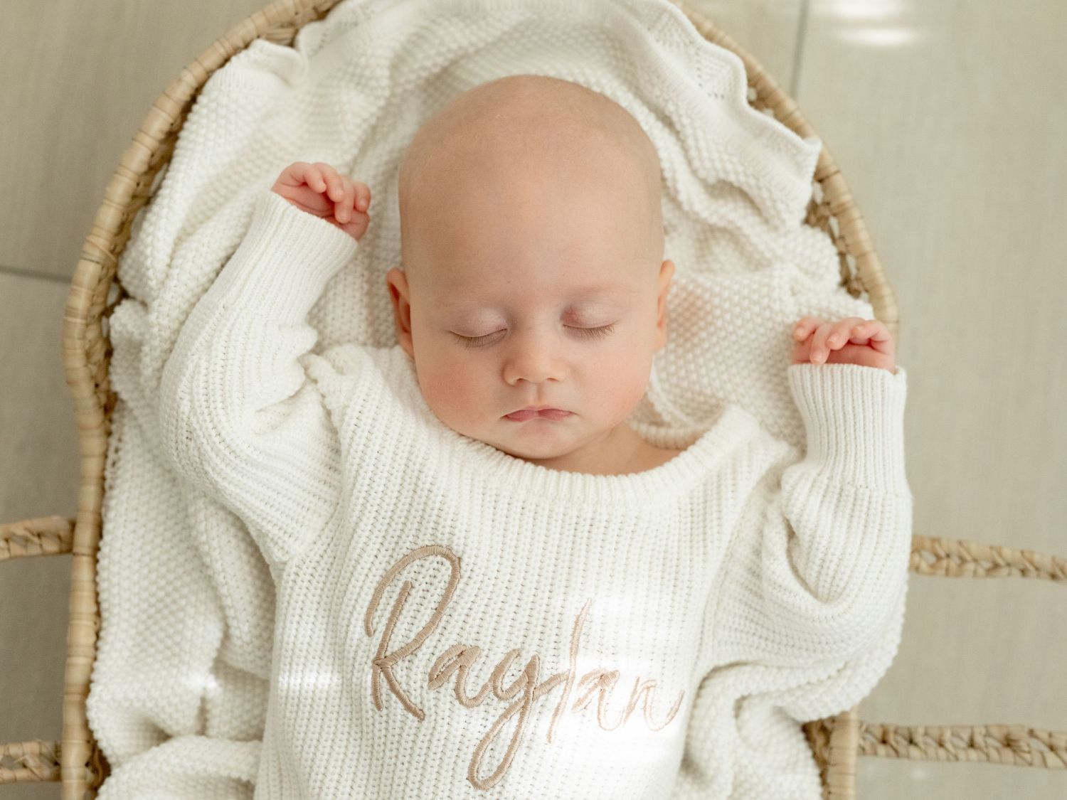 A toddler girl in a white dress and gold headband leans on a white leather pouf ottoman near sheer curtains.