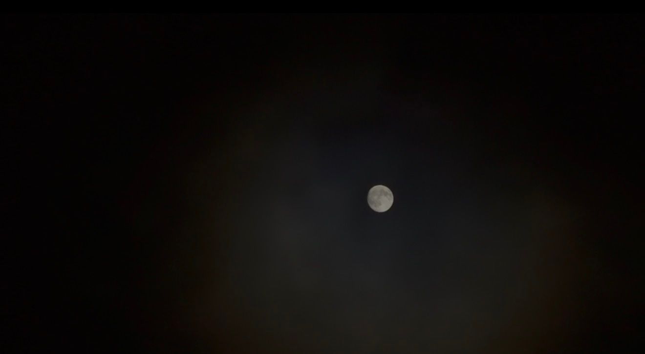 Full moon glowing through dark cloudy night sky, illuminating surrounding clouds with soft white light.