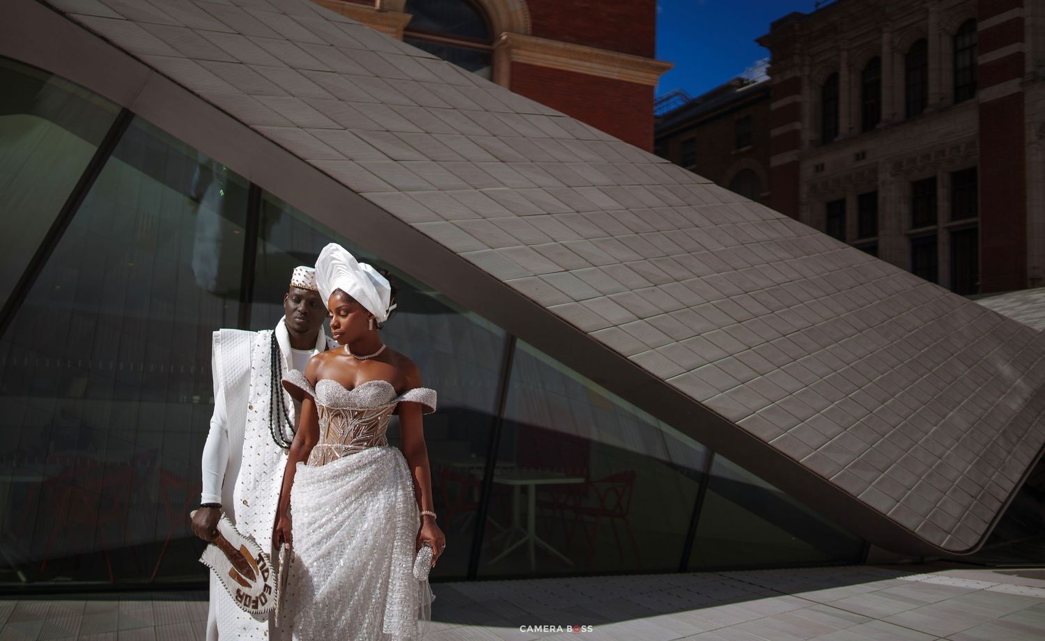 Elegant couple in traditional African wedding attire posing outside a modern geometric building on their wedding day.