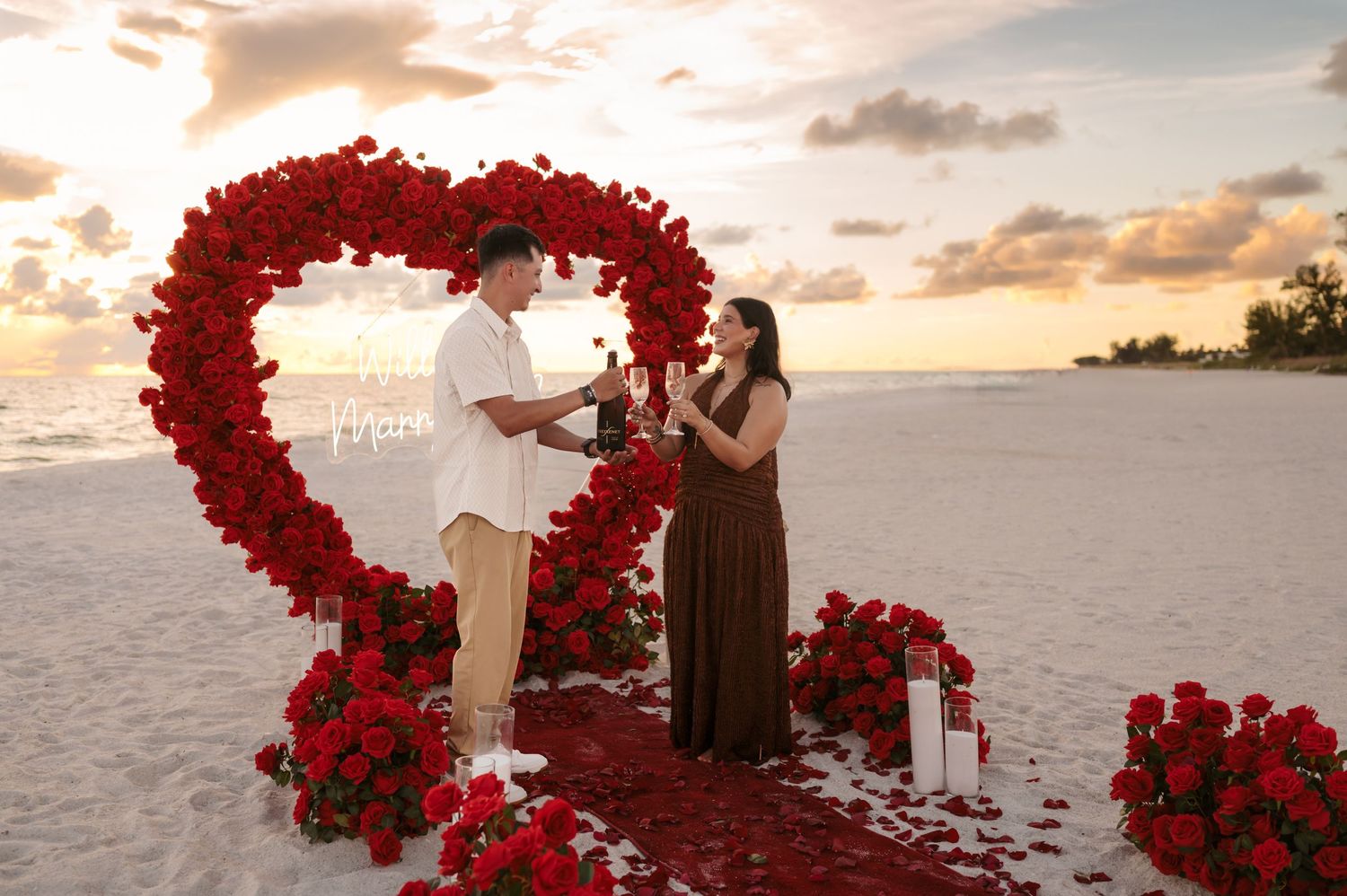 Couple sharing a romantic beach proposal at sunset surrounded by red roses arranged in a heart shape.