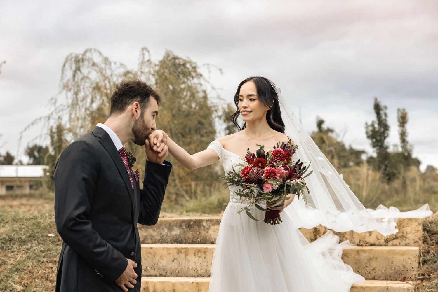 Groom kissing bride's hand outdoors, bride holding red bouquet in white wedding gown, romantic countryside wedding photo.