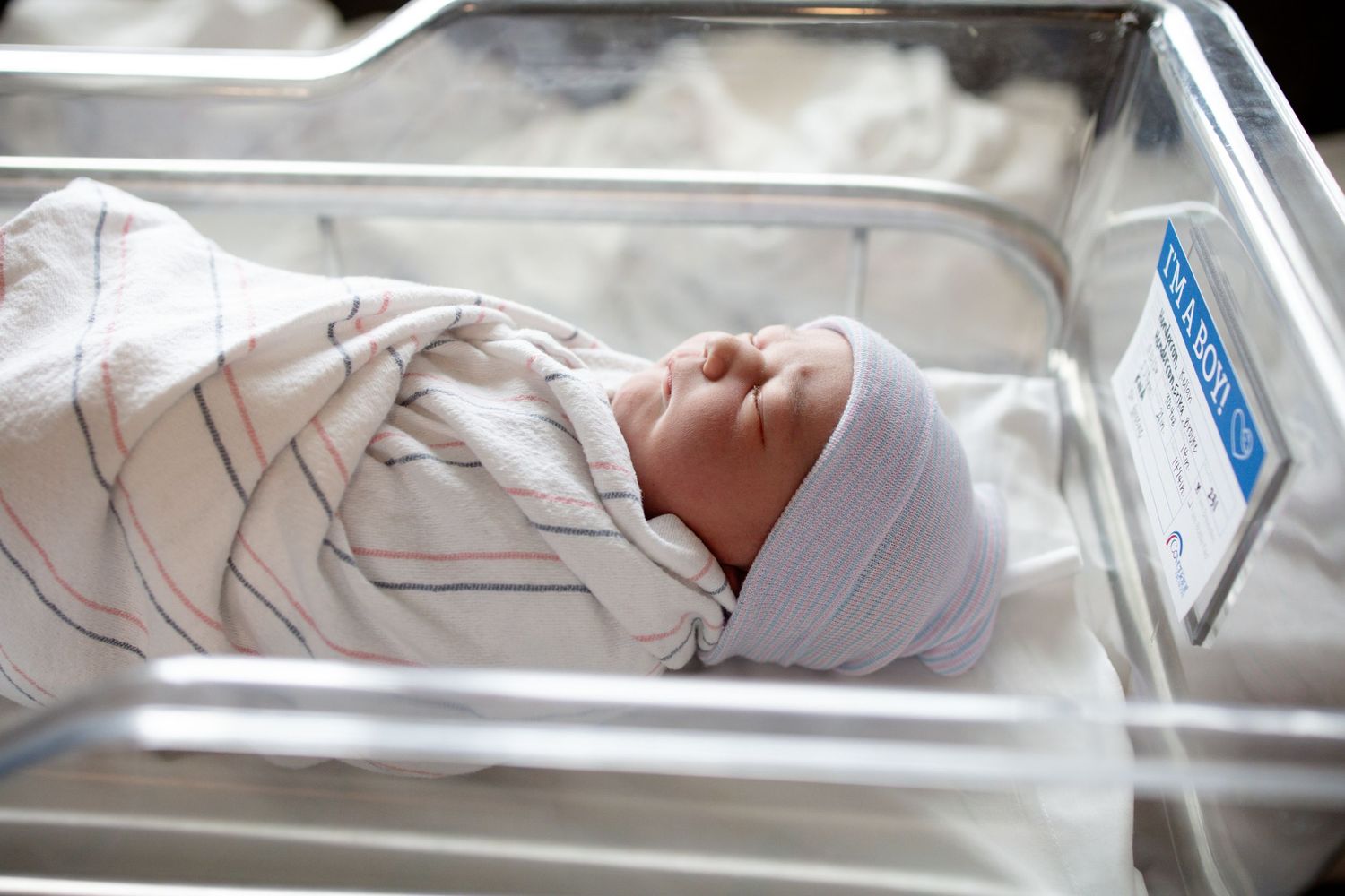 Sleeping newborn baby in a white cap, swaddled in a striped hospital blanket and resting in a clear bassinet with an ID card.