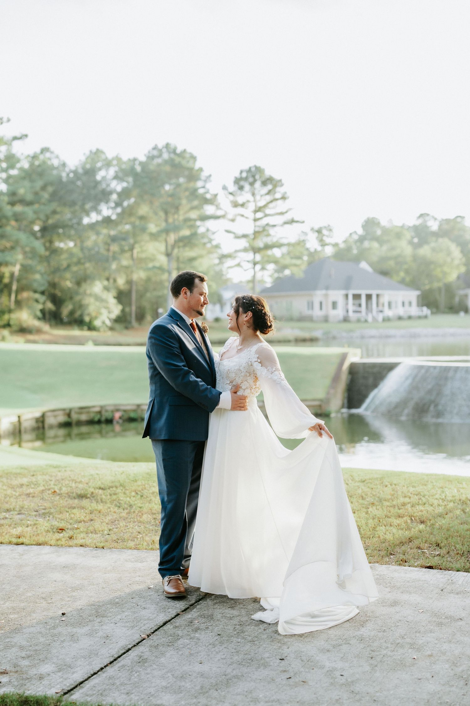 Bride and groom share a romantic moment in wedding attire beside a serene pond with a white estate in the background.