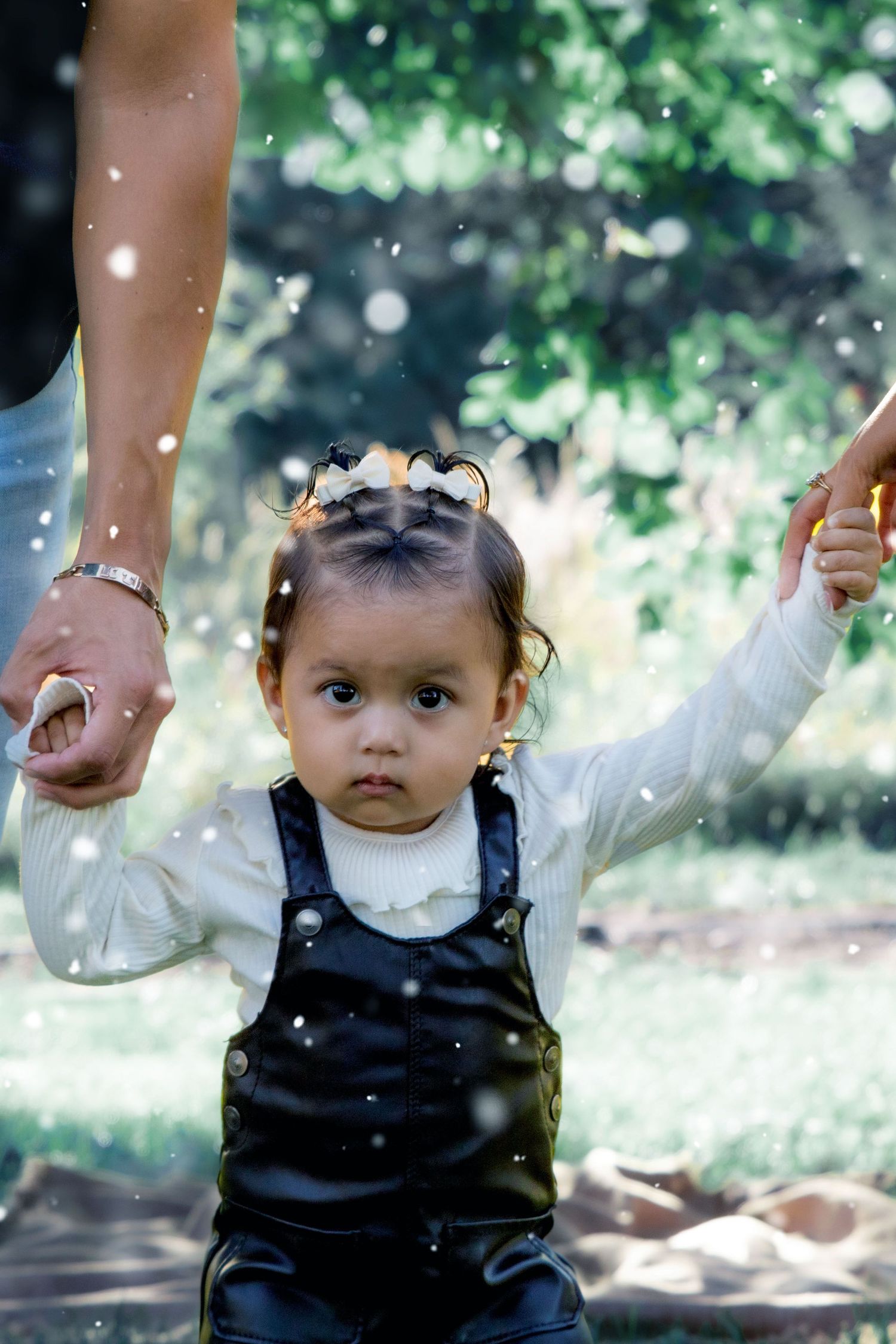 Toddler in black overalls holding hands while walking in a sunlit garden with sparkles.