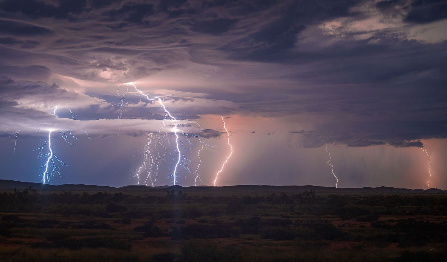 Multiple lightning bolts illuminate a dark stormy sky over a desert landscape at night.