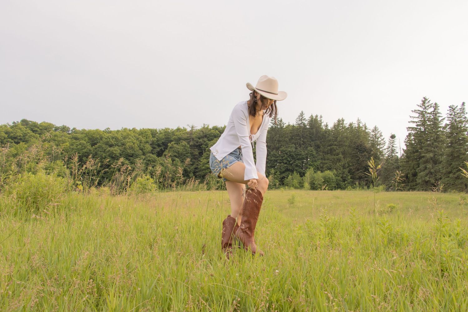 A person in a cowboy hat and boots standing in a grassy field. The person appears to be adjusting their boot while looki