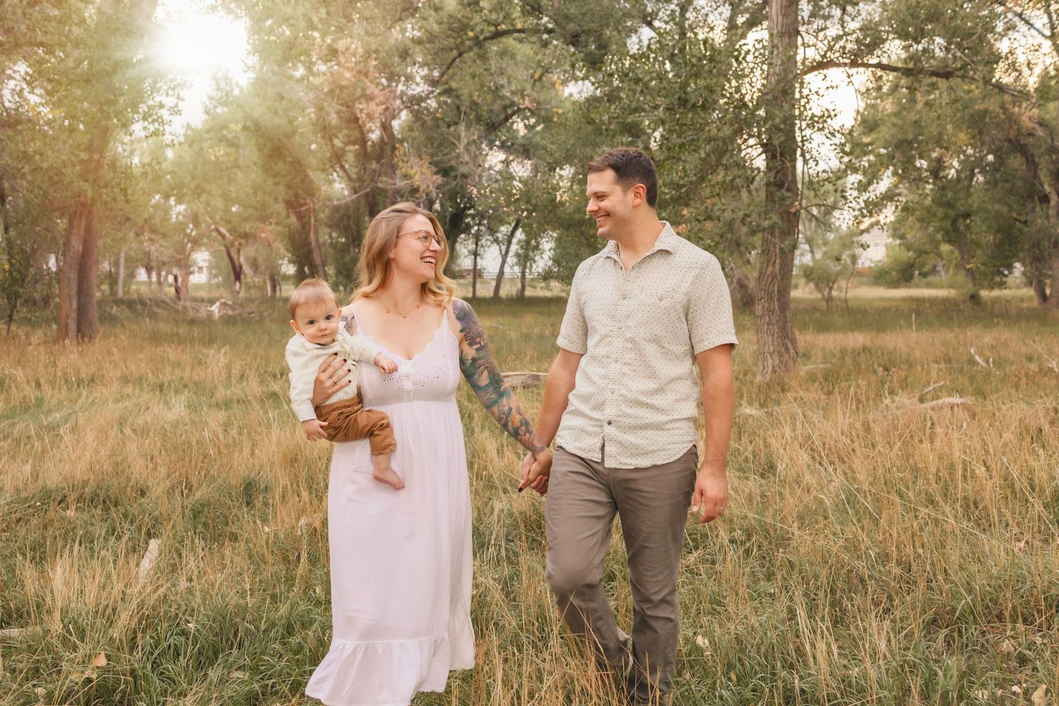 A family walking hand-in-hand through a grassy forest, mom in the middle holding hands with dad and holding baby on hip