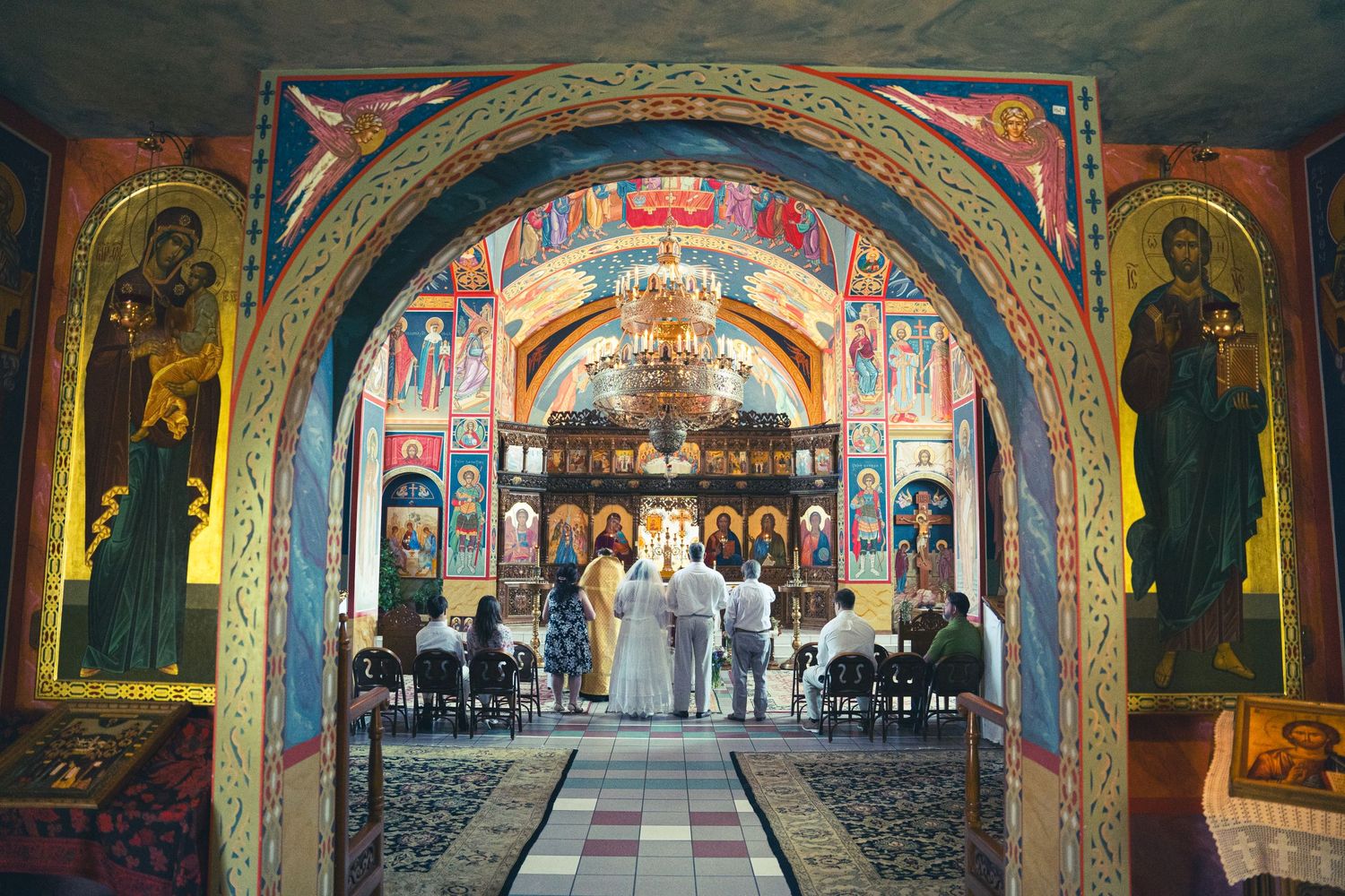 An ornate Orthodox church interior with painted arches and religious iconography during a ceremony.