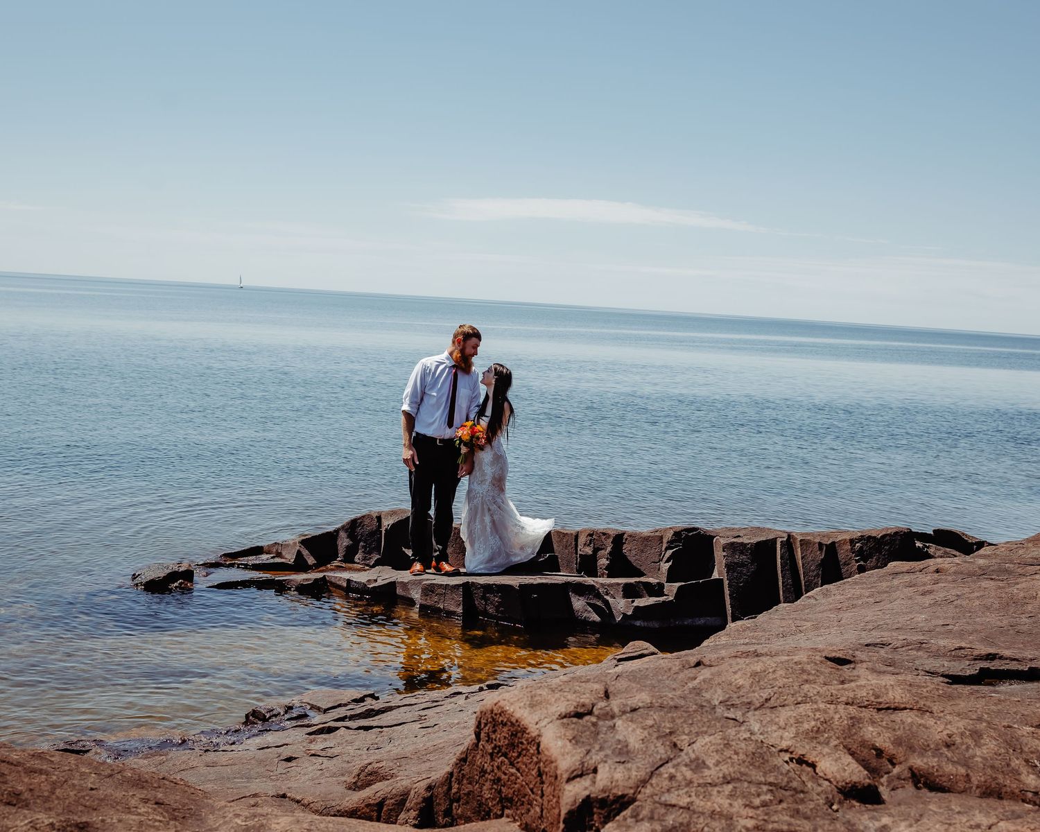 A couple stands together on rocky Lake Superior shores on a bright sunny day.