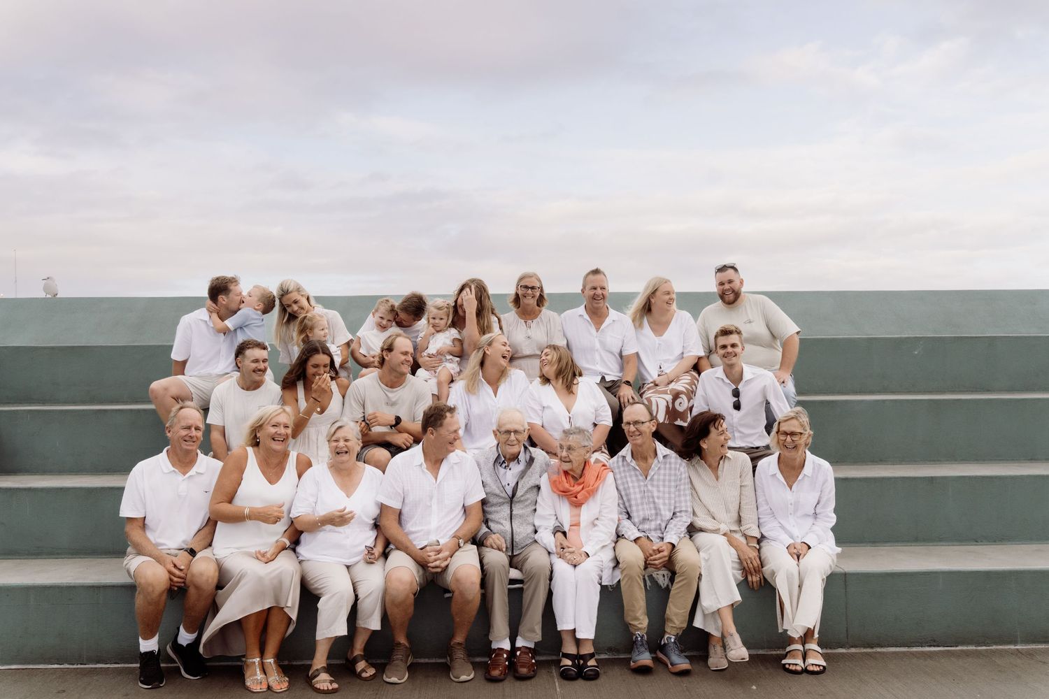 Large family group portrait on outdoor steps near the ocean in white and neutral outfits.