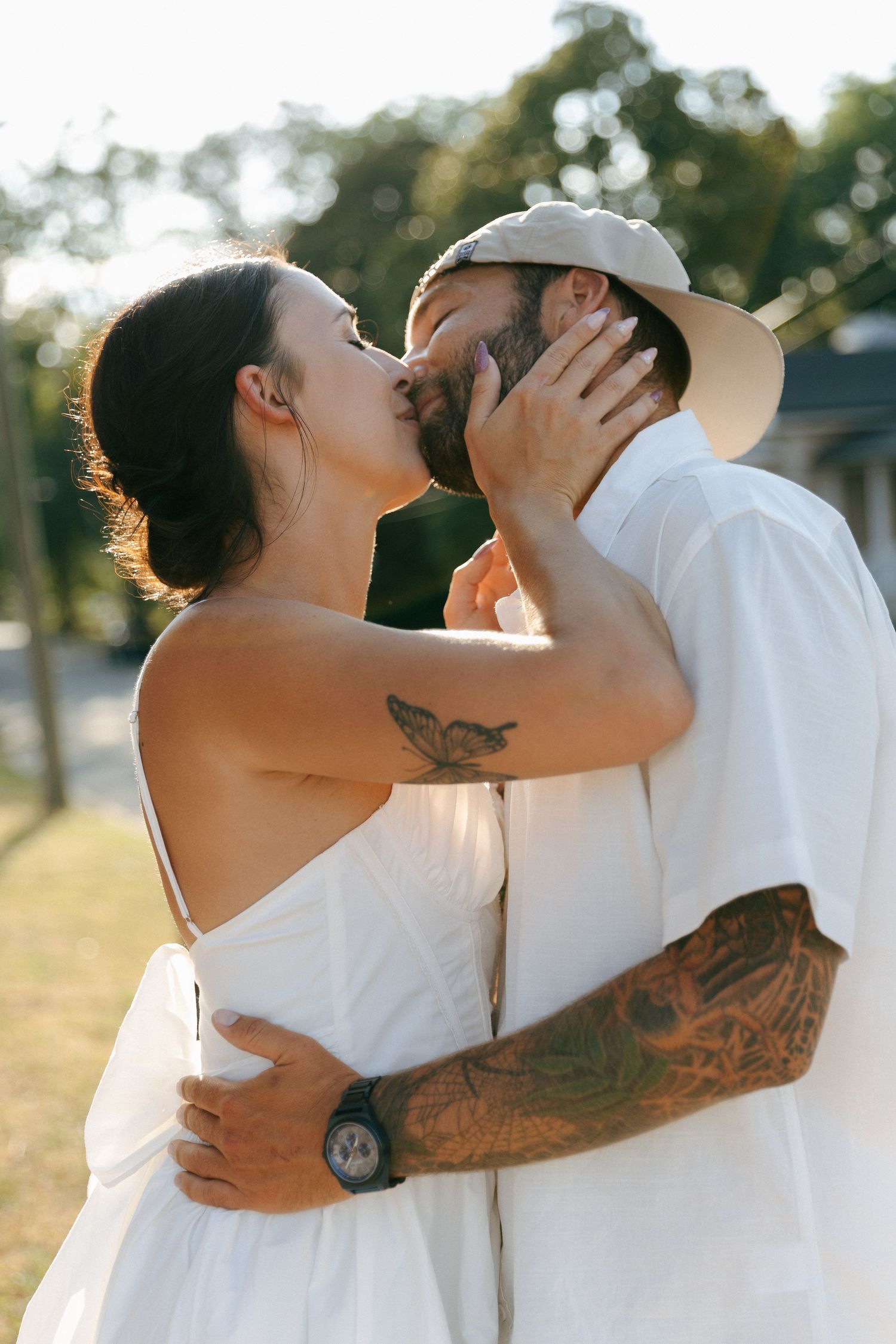 Romantic couple embraces in sunlit outdoor setting wearing white, both with visible tattoos sharing intimate moment in nature.