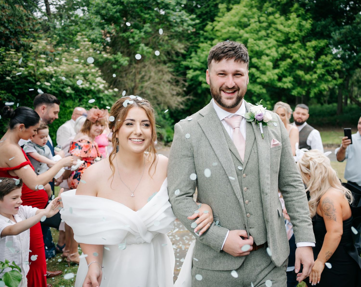 Happy wedding couple walking through confetti shower in outdoor garden celebration with guests.