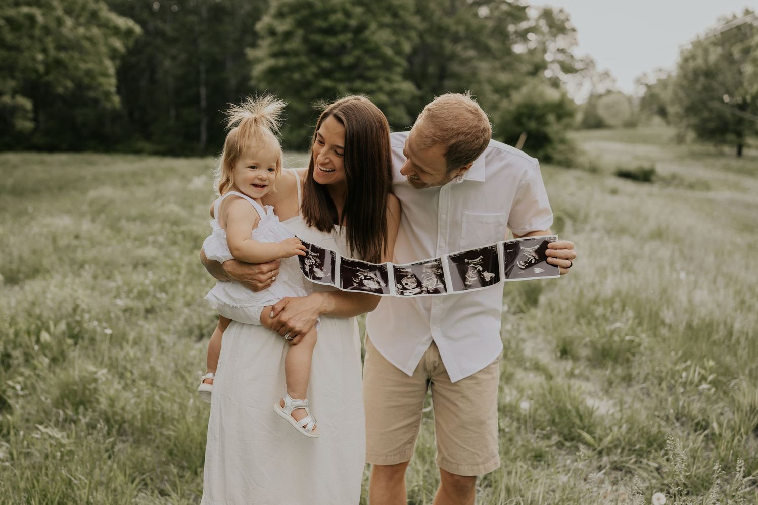 A young family wearing white outfits holds ultrasound photos while posing together in a sunny meadow.