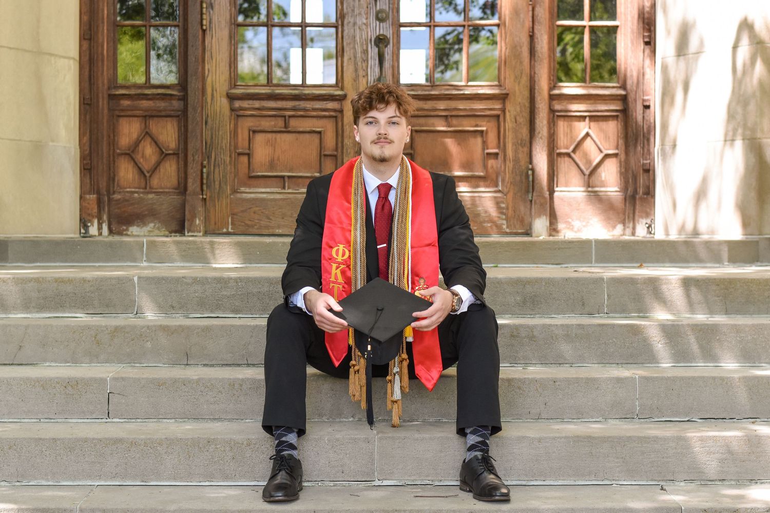 A minister in red religious stole stands against a stone church wall with columns.