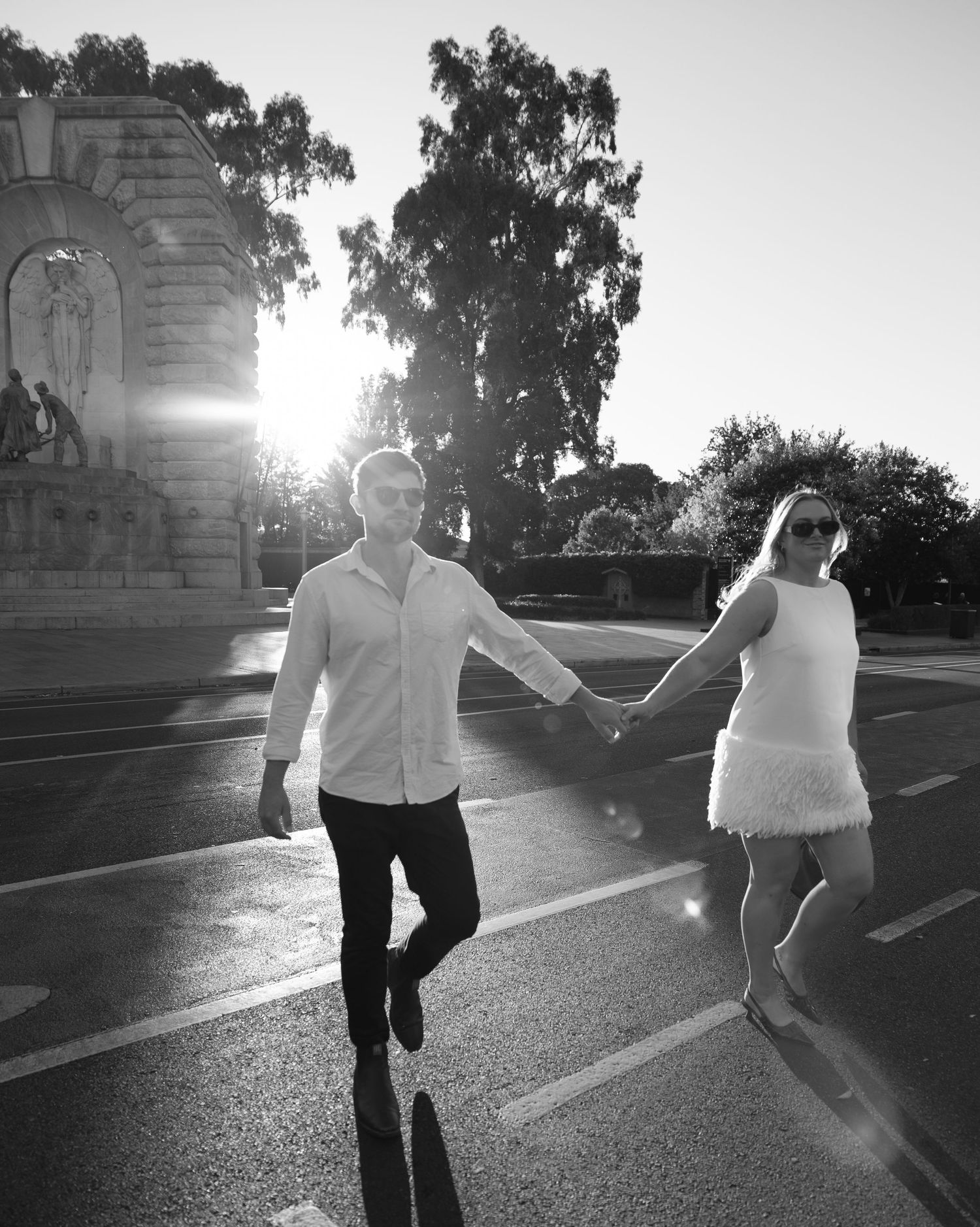 Couple walking together while holding hands in a sun-lit street with ornate architecture in the background.