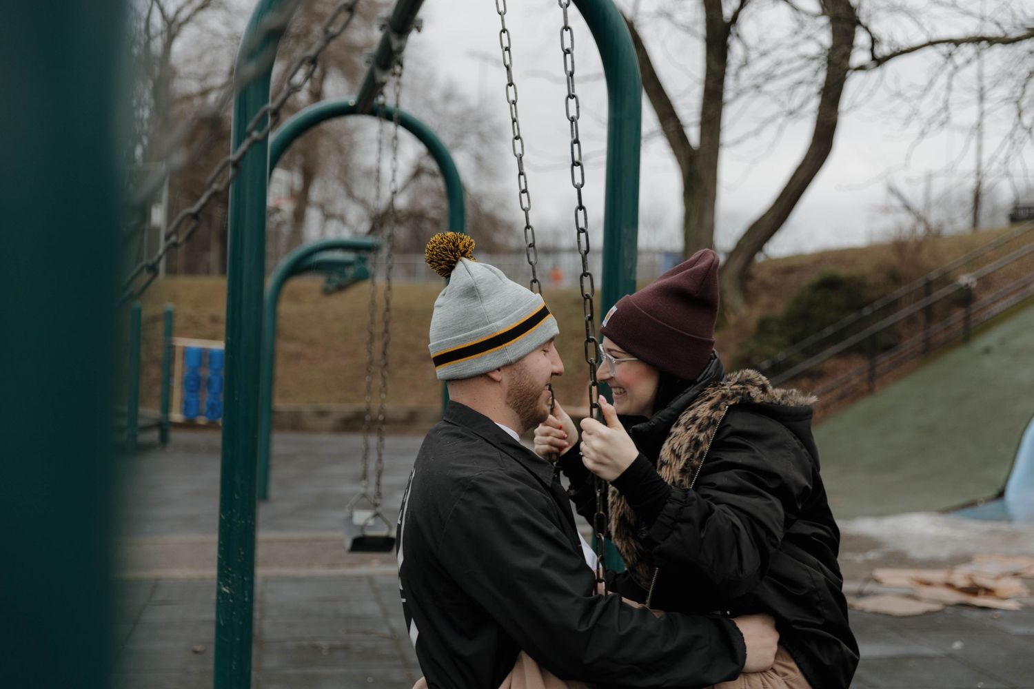 Two people in warm winter hats share a tender moment on a playground swing set on a cold, cloudy day.