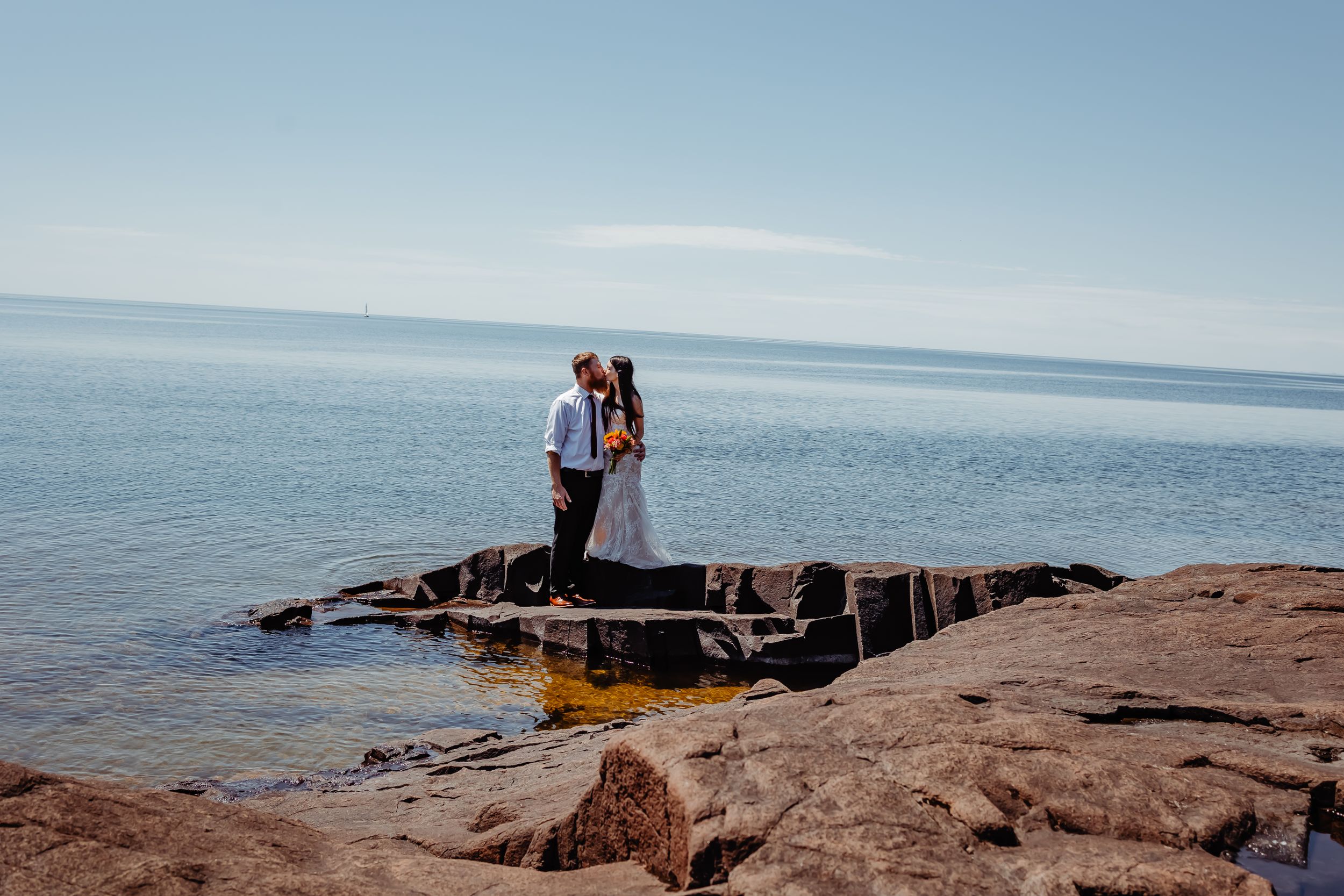 Couple standing on rocky outcrop overlooking vast ocean under blue sky.