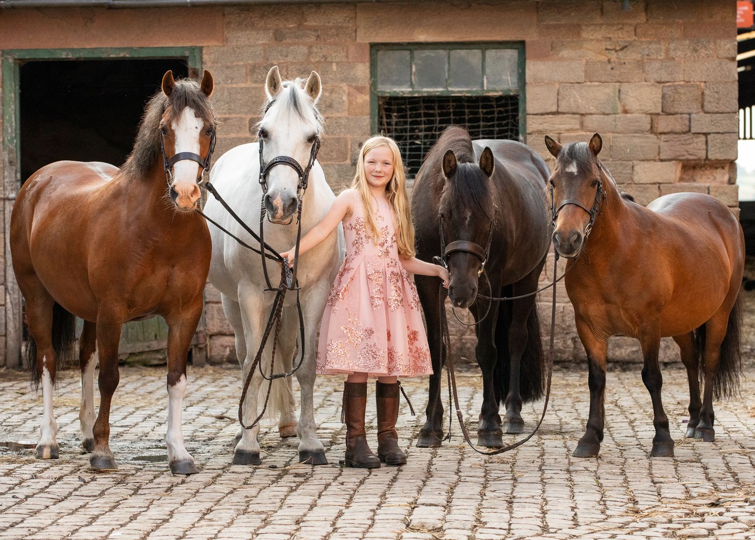 Four beautiful horses stand on cobblestone pavement outside a rustic brick stable with a person in a pink dress between them.