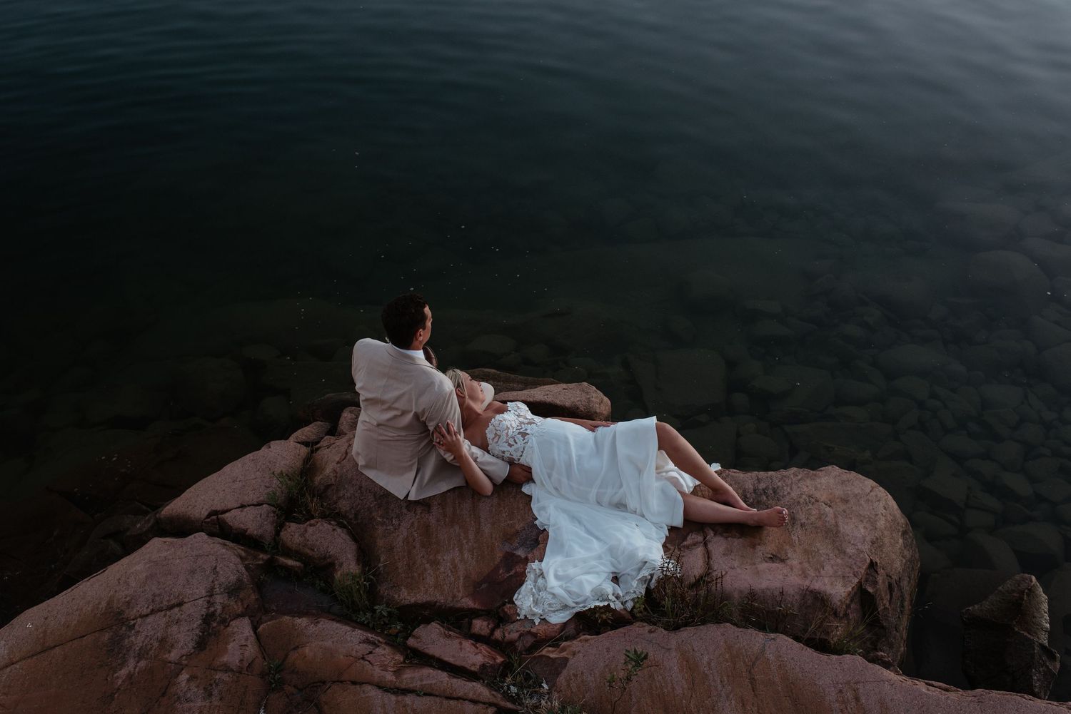 Two people in wedding attire lounge on red rocky cliffs overlooking dark water at dusk.
