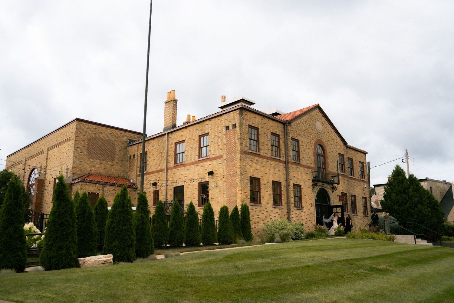 A stately brick mansion with emerald green hedges lines the gently sloping lawn under cloudy skies.
