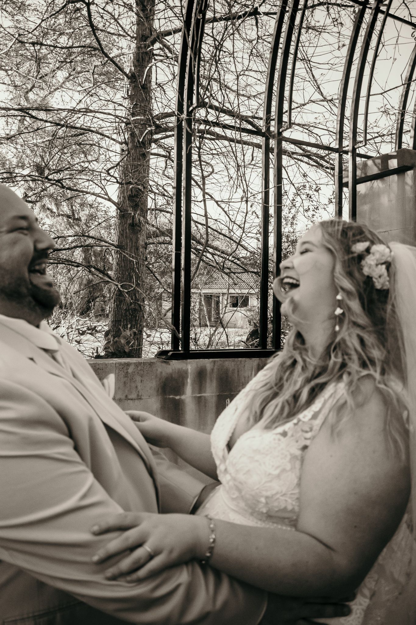 A joyful couple shares a candid laughing moment together beneath bare winter trees in black and white photography.