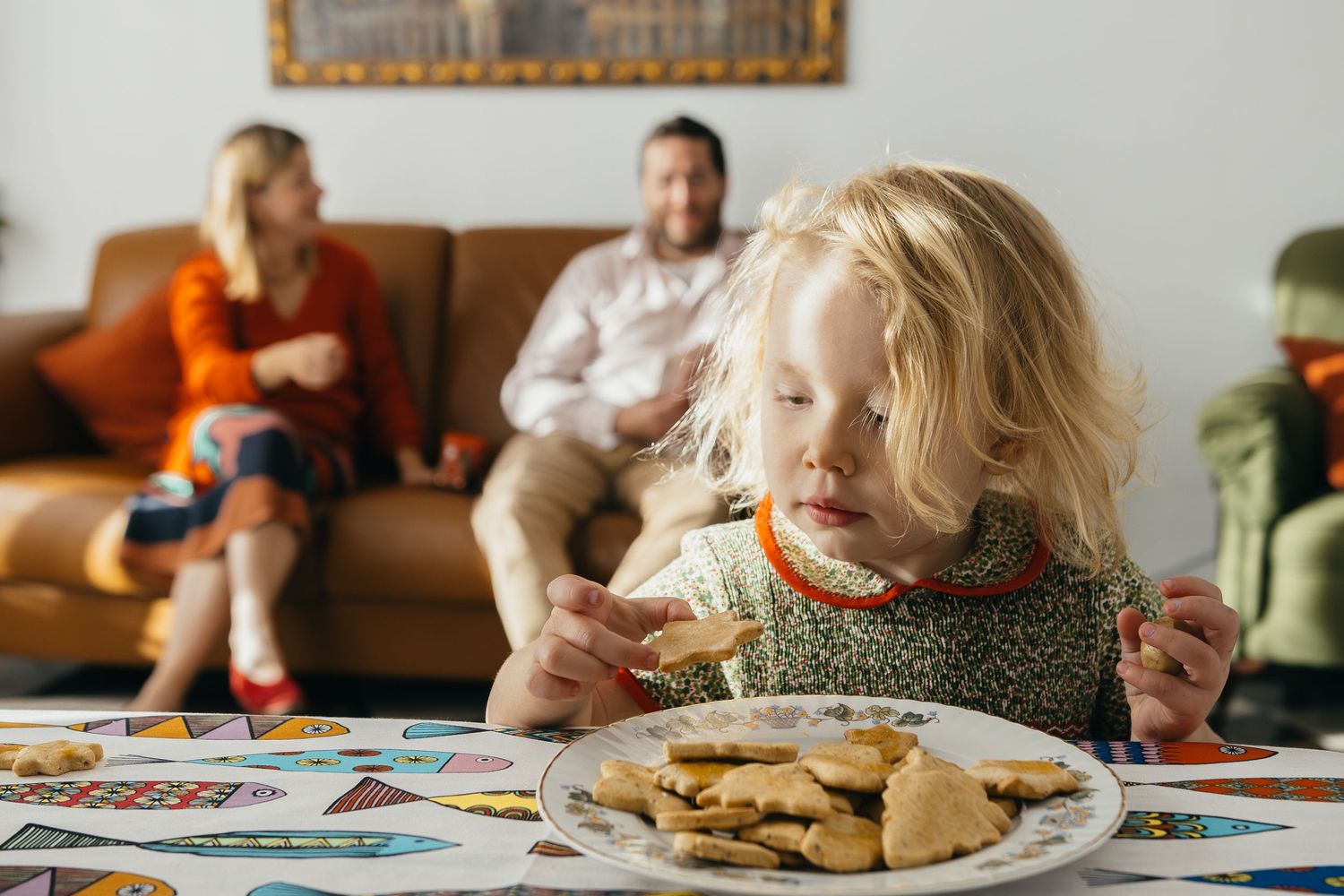 A plate of fresh cookies sits on a colorful table with a couch and people visible in the background.