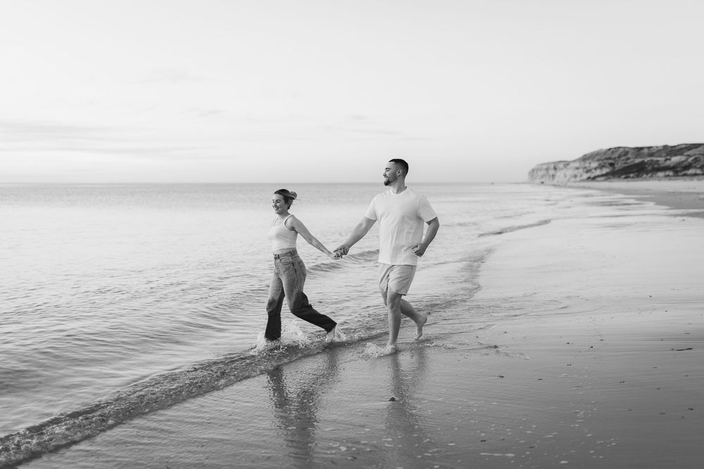 Two people holding hands while walking along the shoreline of a beach at sunset in black and white.