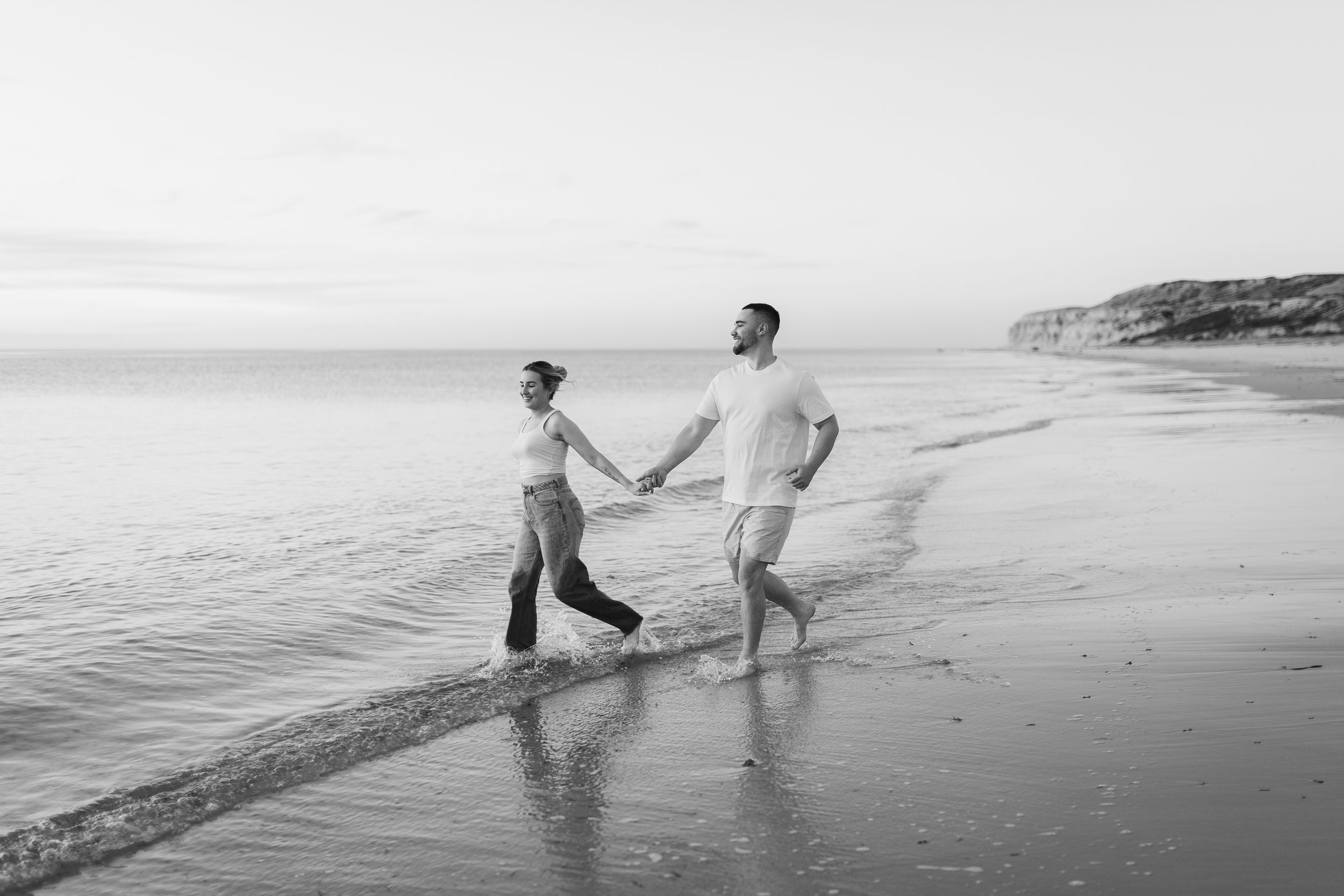 Two people holding hands while walking along the shoreline of a beach at sunset in black and white.