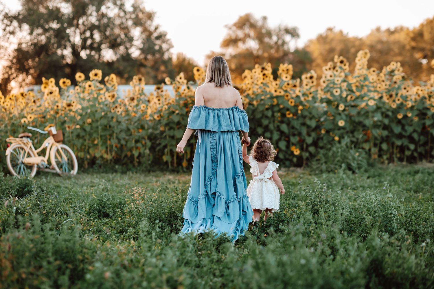 A person in a flowing blue dress and small child walk through a meadow at sunset with sunflowers and a bicycle in background.