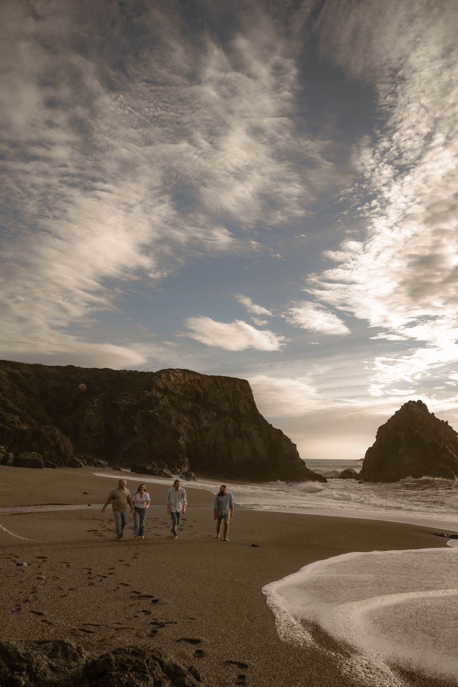 People walking along a dramatic coastal beach with towering cliffs and moody clouds overhead.