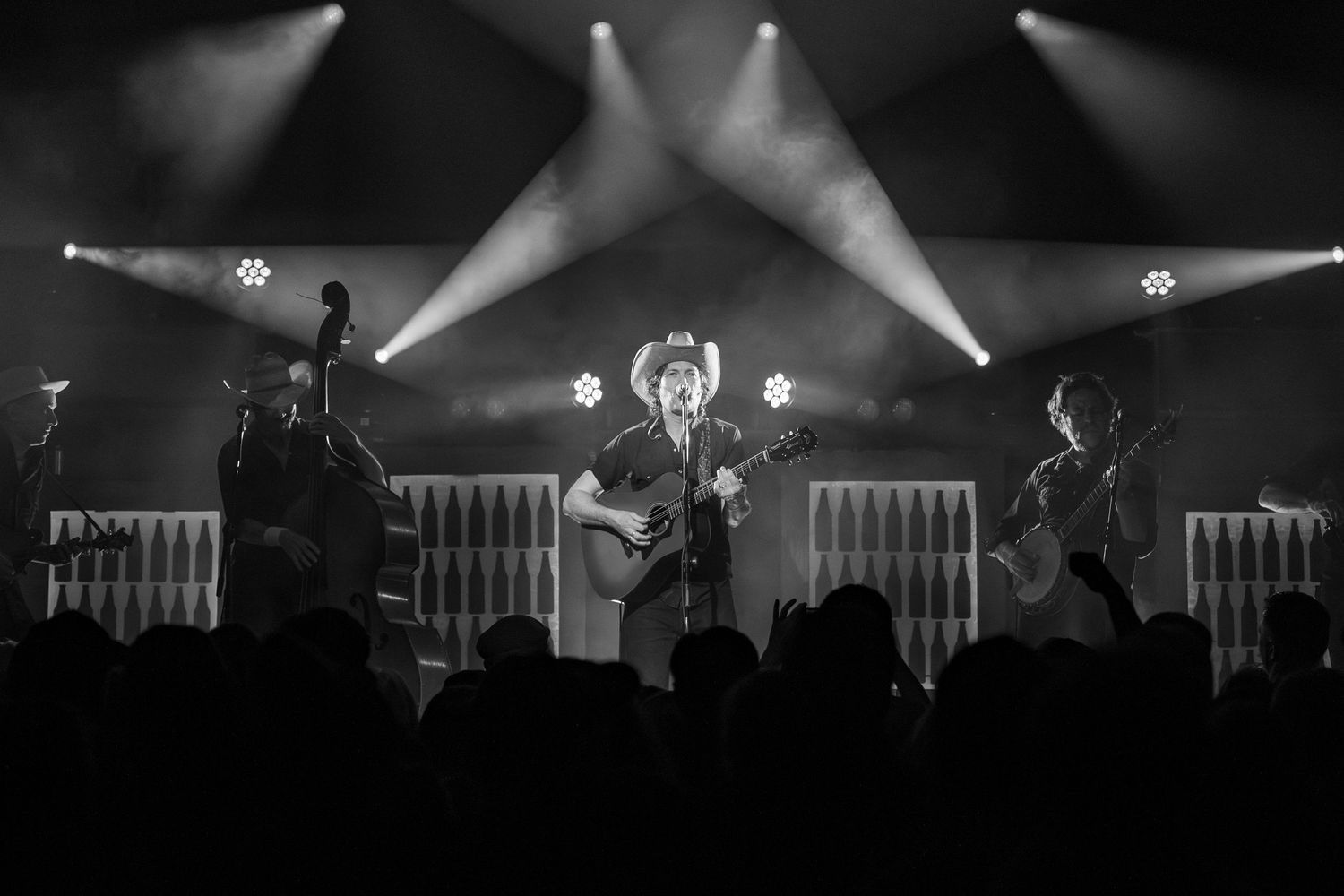 Band performs on concert stage with dramatic spotlight beams and silhouetted crowd in foreground.