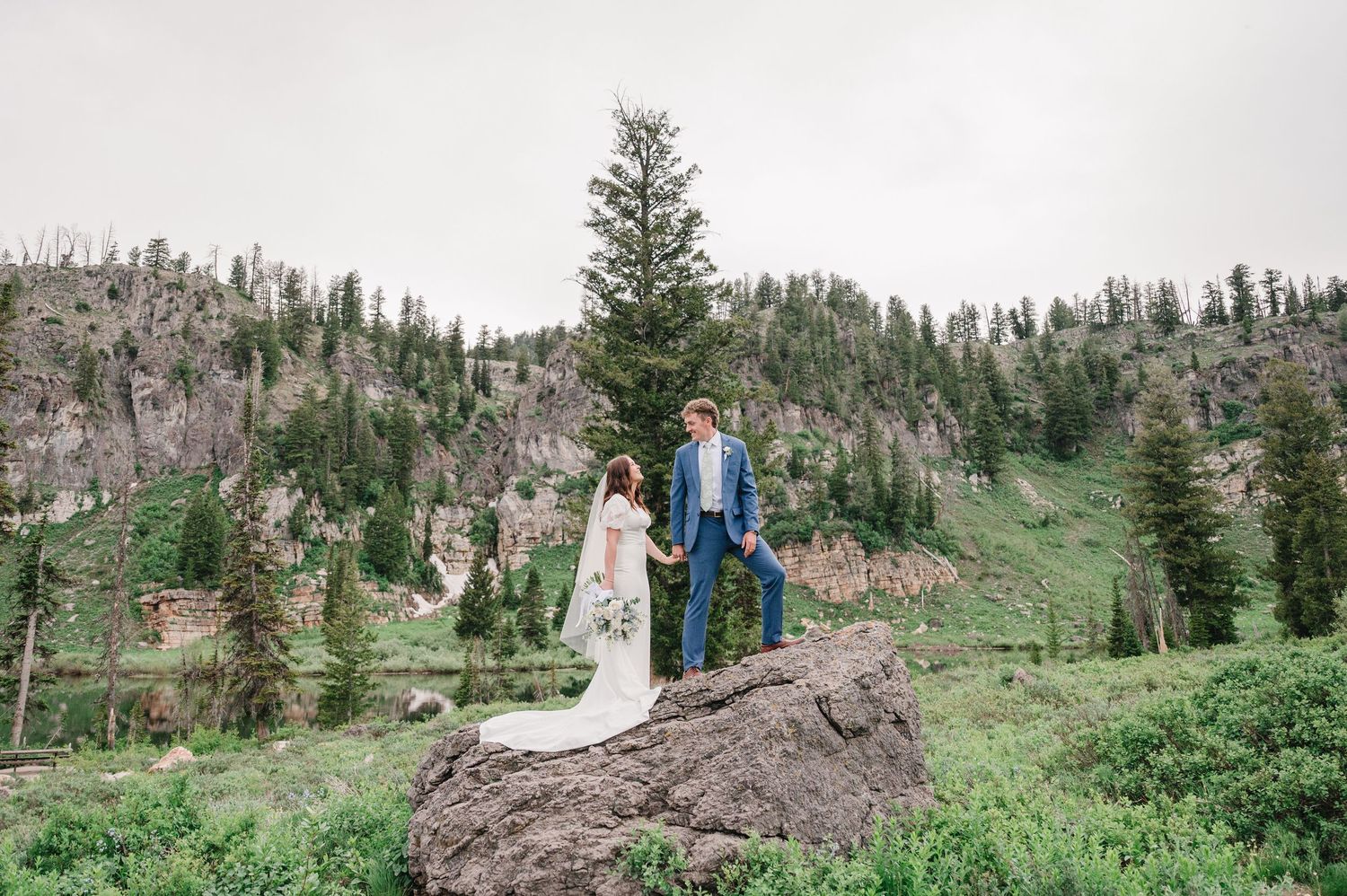 Two figures stand on a rocky outcrop surrounded by evergreen trees and mountain slopes in a scenic wilderness setting.