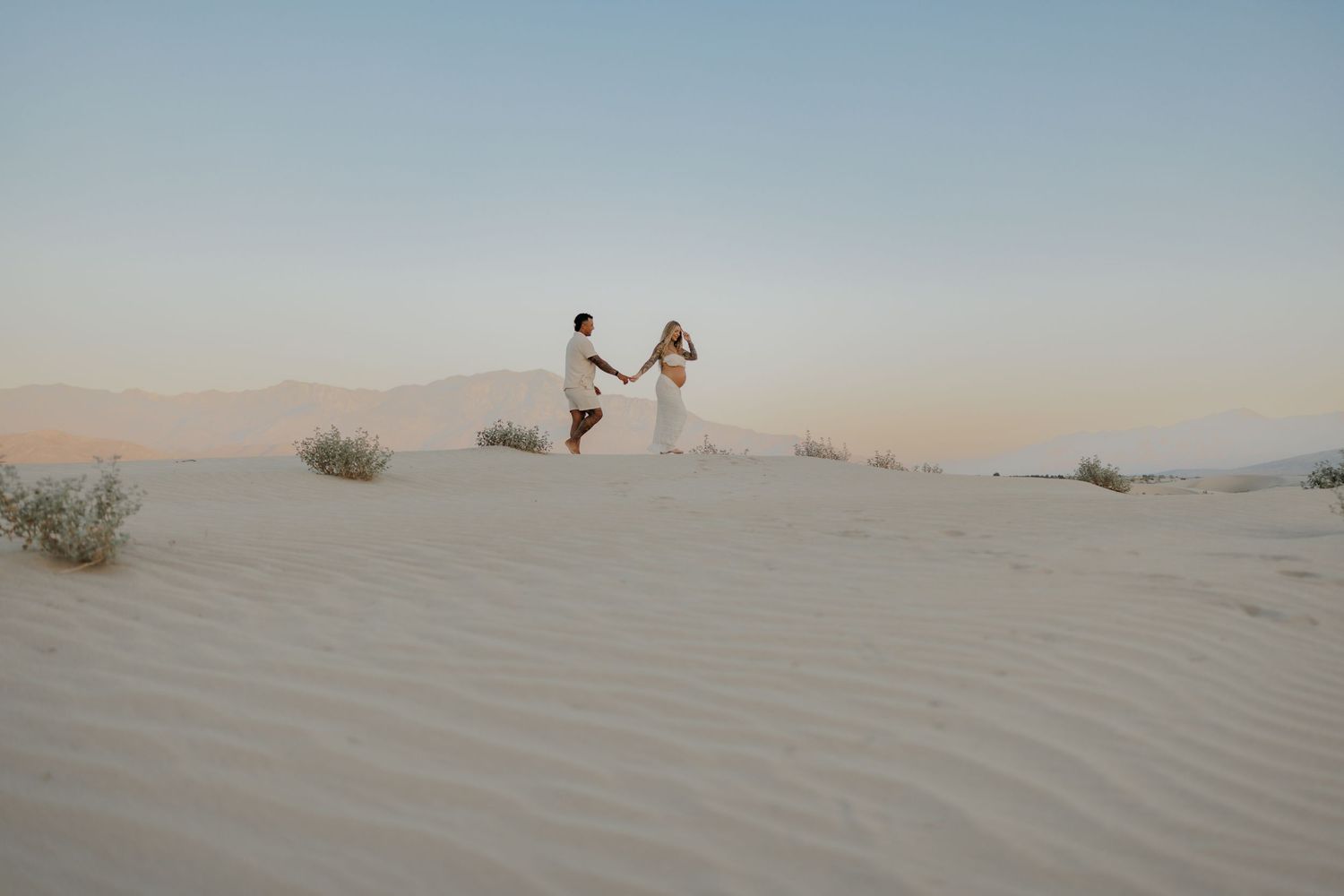 Couple holding hands while walking on sandy dunes during sunset.