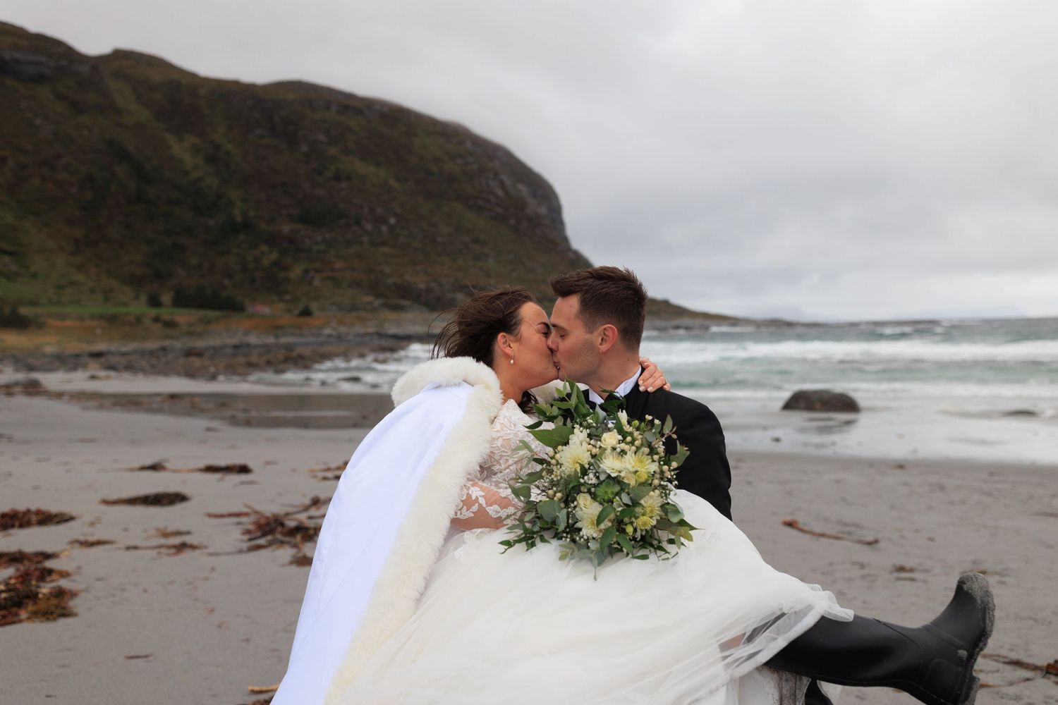 A wedding couple shares a kiss on a rocky beach with crashing waves and mountains in the background.