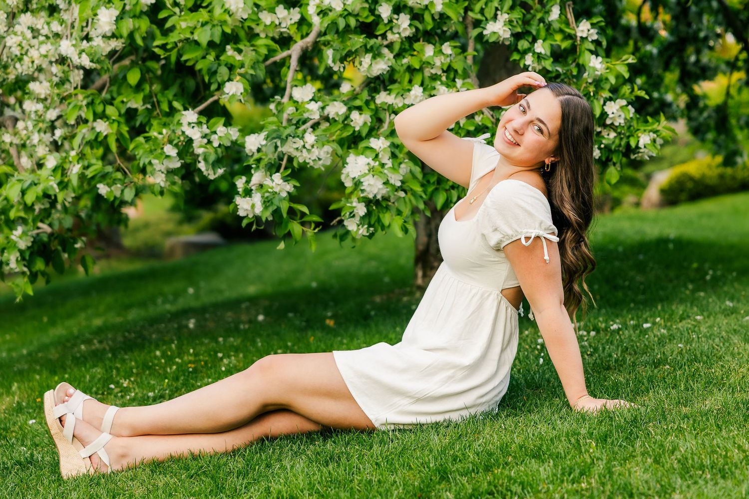 Woman in white summer dress relaxing on grass beneath blooming spring tree branches in garden setting.