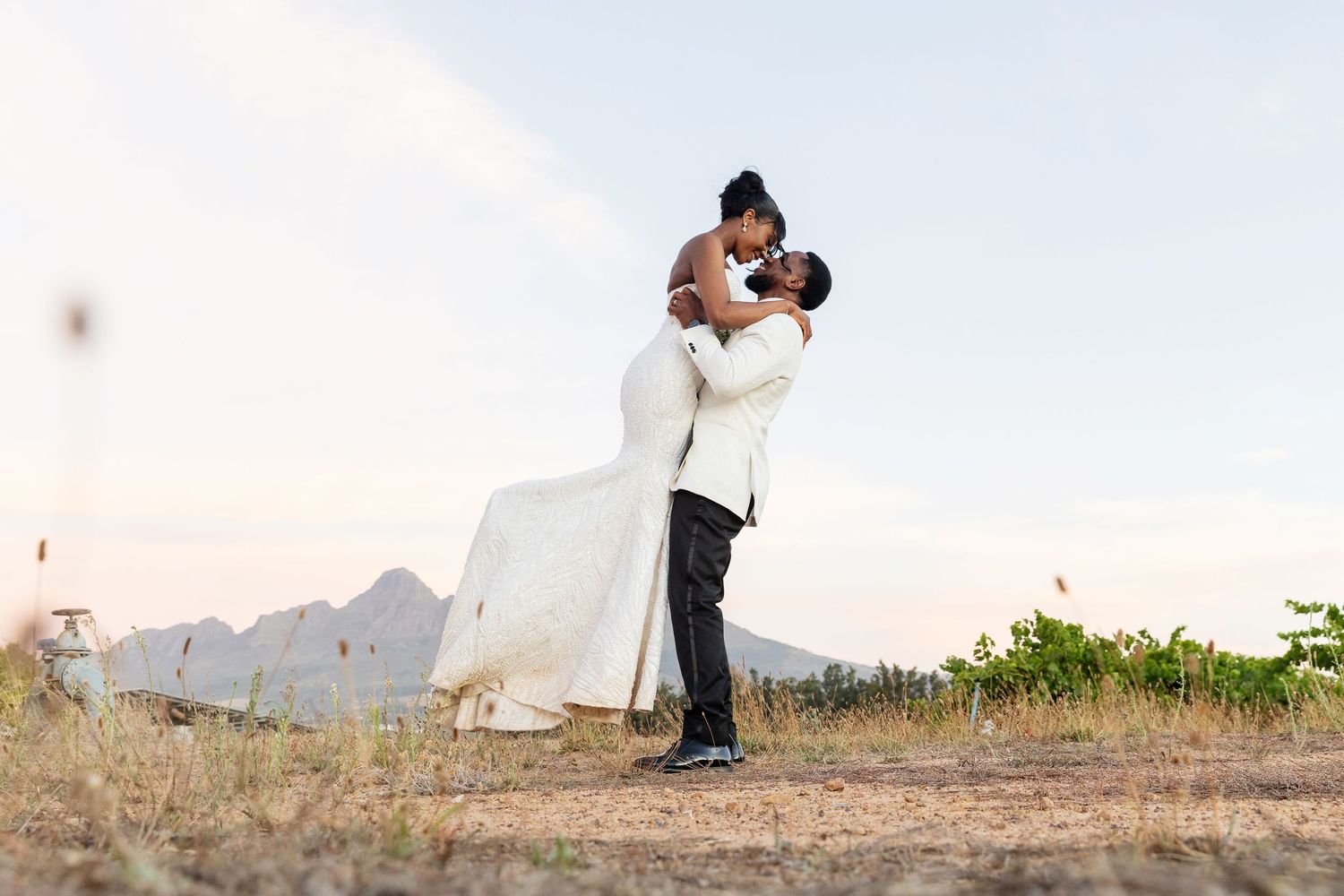 A couple shares a romantic embrace in a white wedding dress and suit against a mountainous backdrop at golden hour.