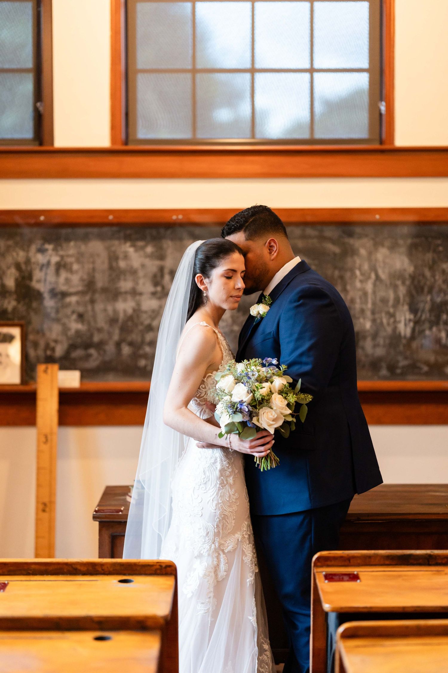 A couple in wedding attire embrace on a lush green lawn surrounded by mature trees in an outdoor garden setting.