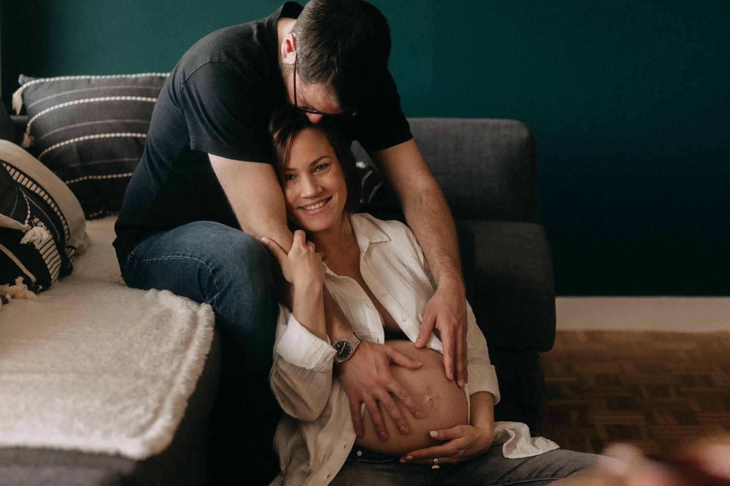 A couple shares an intimate moment while relaxing together on a grey couch in a dimly lit room.