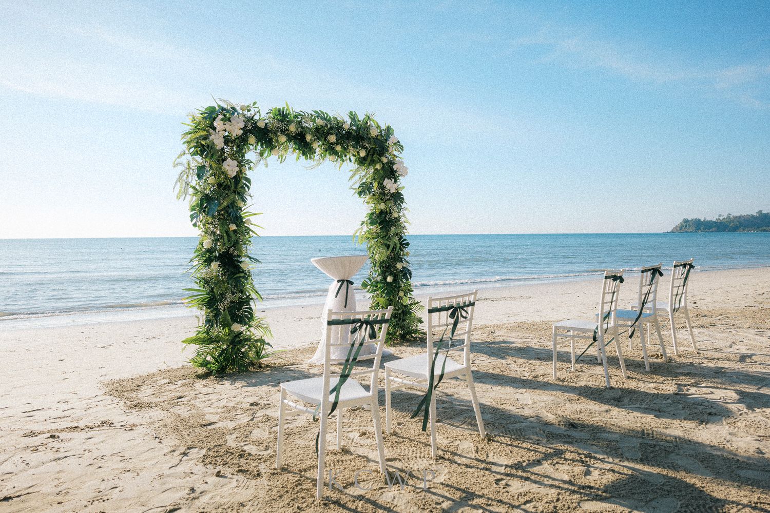A floral wedding arch and white chairs set up for a beach ceremony with ocean waves in the background.