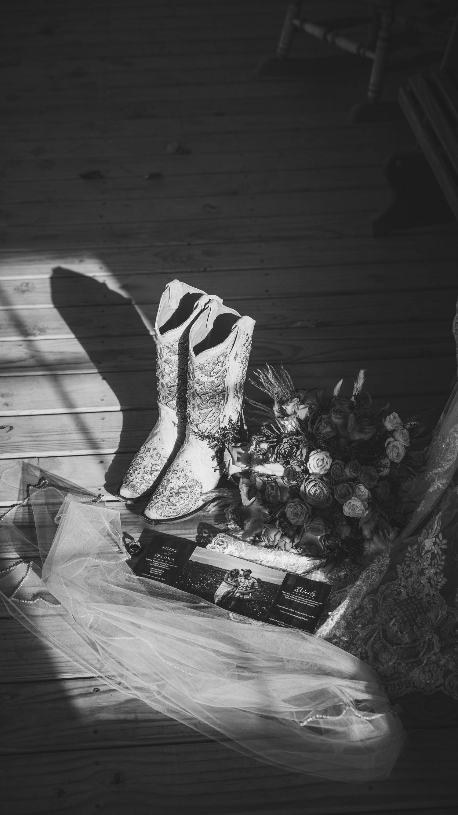 Black and white still life of cowboy boots with roses and veil in dramatic lighting.