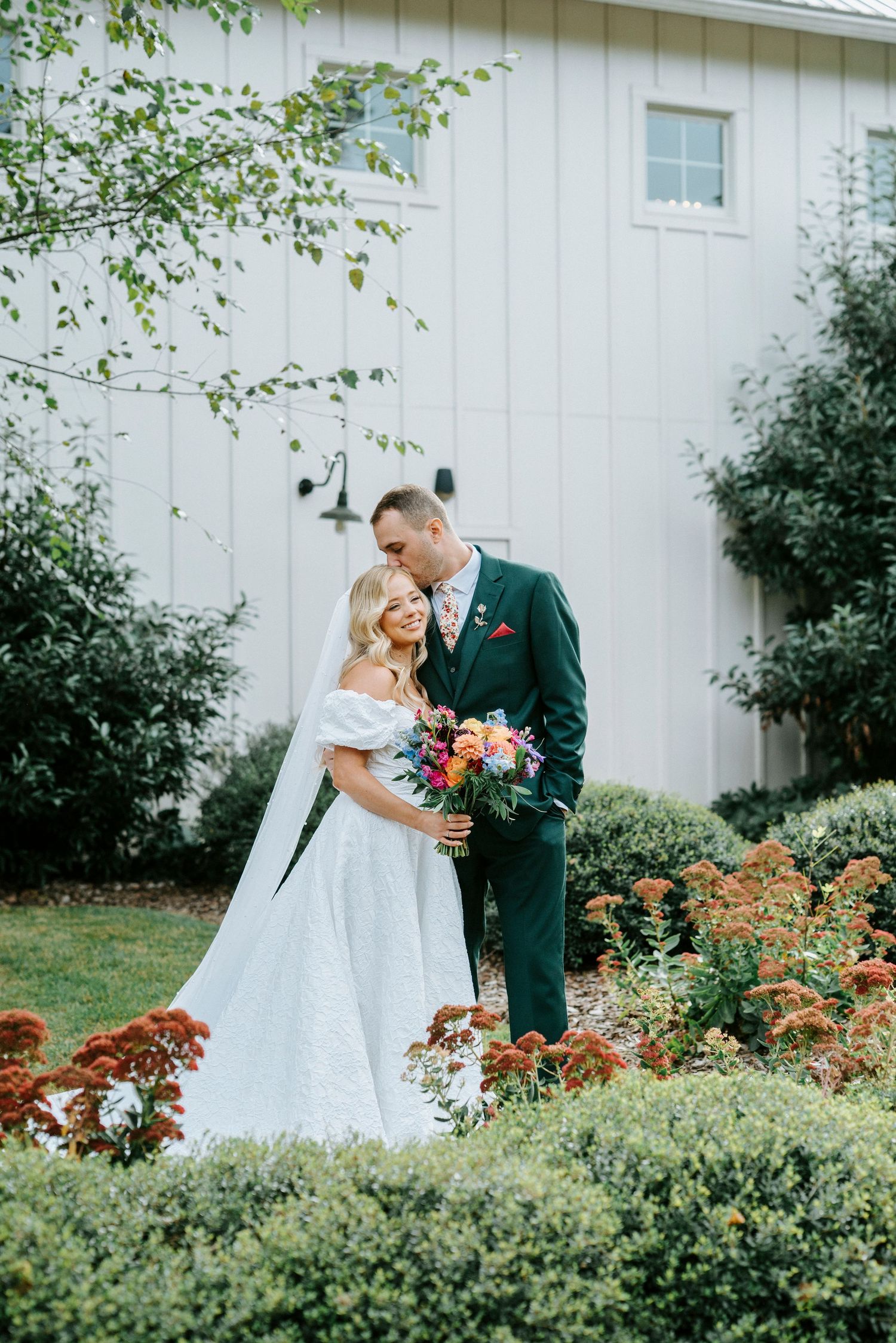 A couple in wedding attire share an intimate moment in a garden with white barn backdrop and colorful florals.
