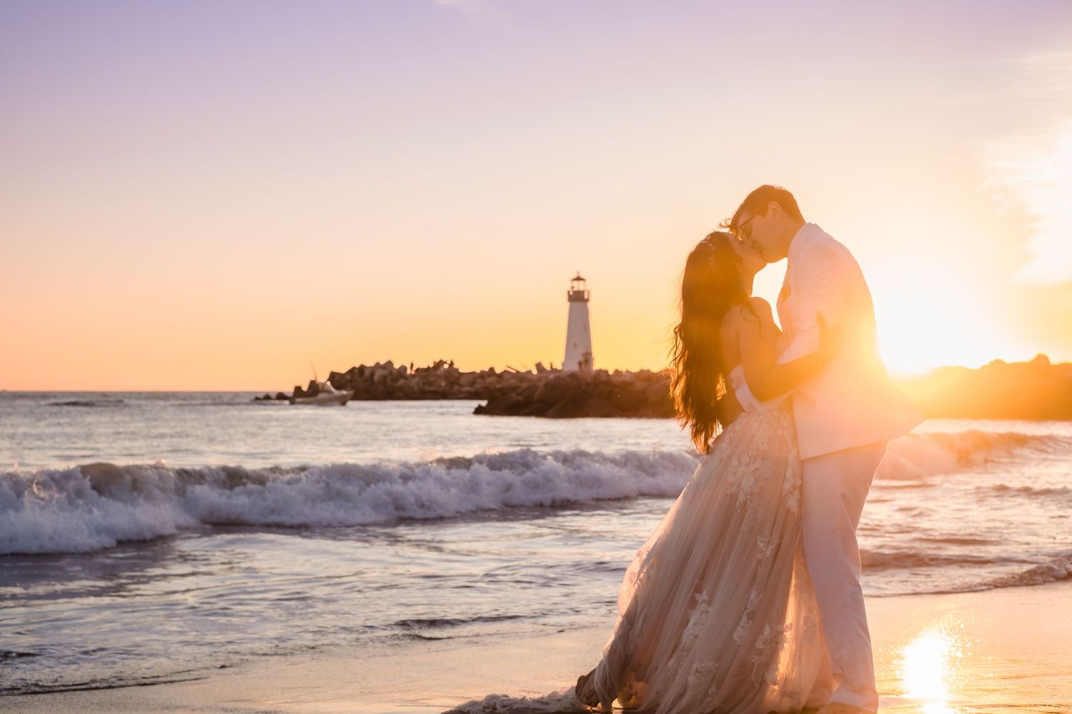 A bride and groom embraces on the beach at sunset with waves crashing and Walton lighthouse in background. On a beautiful Santa Cruz beach.